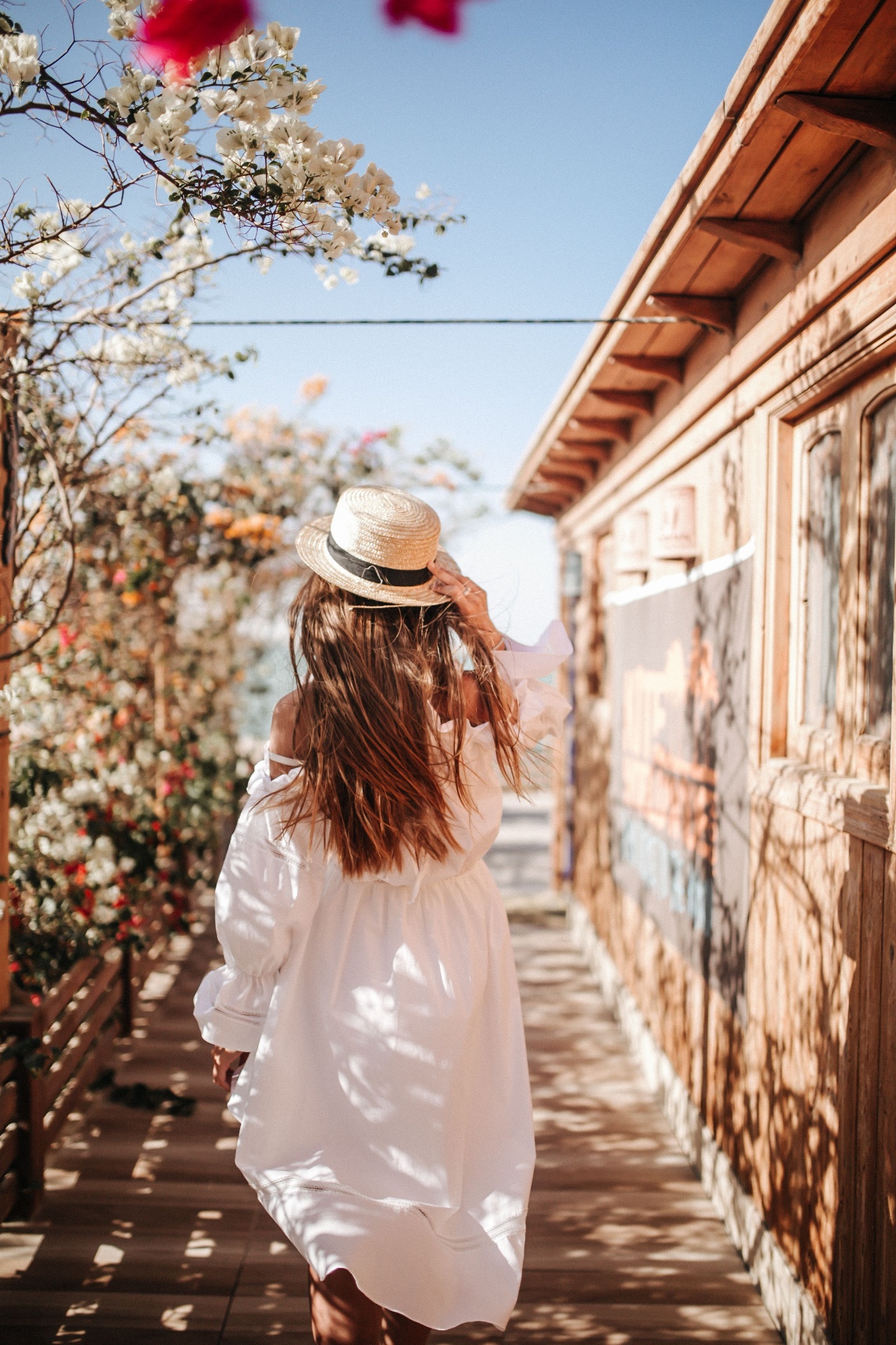 Femme en robe blanche portant un chapeau de paille dans une ruelle fleurie